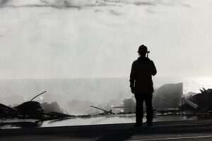 Silhouette of firefighter on roof of destroyed Pacific Palisades home after recent wildfire